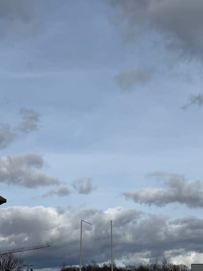 The image depicts an outdoor scene in Oberhausbergen, France, under a partially cloudy sky. The upper two-thirds of the frame show a light blue sky interspersed with various grey, white, and darker cumulus clouds. The lower third of the image primarily features an obscured horizon line with a dense band of dark, leafless trees or shrubs.
In the mid-ground, two tall, slender, light-colored poles stand vertically. The left pole has a horizontal extension projecting to its left from the top. The right pole is a straight vertical column. To the far left, a dark, angled roofline of a building or structure is visible. Below this roofline and above the tree line, the red boom of a construction crane extends diagonally upwards from left to center. A light-colored, flat-topped structure is partially visible on the far right, just above the tree line. No people or active interactions are present in the scene.