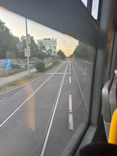 The image captures a view from inside a tram moving along a set of tram tracks in Leipzig, Germany. The tram is on a paved road, with the tracks laid within the road surface. On the left side of the tracks, there is a sidewalk, followed by a grassy area with trees and some construction materials, including large concrete pipes and stacked concrete slabs. A traffic light is visible on a pole. In the background, a white building resembling a castle or a manor house with crenellated battlements stands amidst trees. The sky is clear and light, suggesting either early morning or late afternoon. The perspective suggests the tram is moving forward, with the tracks receding into the distance. The window of the tram is reflecting the environment.
