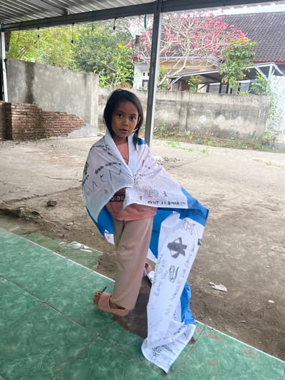 In Desa Batu Layar, Indonesia, a young girl stands under a metal roof structure, directly facing the camera. She is wearing a light orange long-sleeve top, light brown pants, and pink open-toed slide sandals. A white fabric with a central horizontal blue stripe is draped over her shoulders, extending down her sides. The fabric displays various handwritten messages and drawings in black marker, including the word "Israel" on the upper white section, and "Love Your Neighbor," an airplane graphic, and footprints on the lower white section. The immediate foreground consists of a green tiled step or platform on the left and a dirt ground surface on the right. The background features a textured concrete wall, partially exposing red bricks on the lower left, beyond which lush green trees, a flowering tree with pink blossoms, and a house with a dark tiled roof and light-colored walls are visible. The setting is an outdoor, partially covered area.