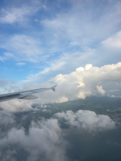 An aerial view of the coast near Ban Tai, Thailand, is captured from an airplane window. The image shows the plane's wing extending into the frame from the left, slicing through a layer of fluffy white clouds that partially obscure the landscape below. The land appears mountainous and covered in lush green vegetation, with glimpses of a dark blue body of water and a coastal line. The sky above is a vibrant blue, dotted with scattered white clouds, some of which are illuminated by sunlight, creating a dramatic contrast with the shaded areas. The scene suggests a flight over a tropical coastal region, likely during the daytime given the bright sky and illuminated clouds.