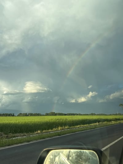 The image shows a view from inside a vehicle, likely a car, moving along a road. The road is paved with a white line marking the edge. To the right of the road is a green field or grassy area with a line of trees along the edge. In the sky, there are clouds, and a faint rainbow is visible. The rearview mirror of the car is visible in the right corner of the frame. The photograph was taken in the Gabarevo, Bulgaria, in the daytime.