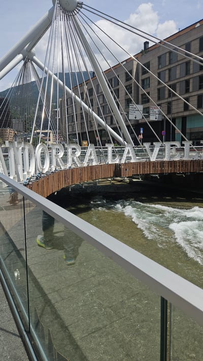 The image captures a detailed view of a modern pedestrian bridge in Andorra la Vella, Andorra, spanning a river with noticeable currents. The bridge features a distinctive white, cable-stayed structure, with numerous taut cables radiating from central hubs. Large, three-dimensional white letters spell out "ANDORRA LA VELLA" along the side of the bridge, positioned above a wooden paneling that forms part of the bridge's underside. The river beneath the bridge flows from left to right, exhibiting choppy waters and whitecaps. On the far bank, multi-story buildings with numerous windows line the river, backed by distant mountains under a partially cloudy blue sky. Several road signs are visible on the far side of the river, one indicating "ORDINO LA MASSANA" with directional arrows. In the immediate foreground, a glass and aluminum railing delineates the bridge's walkway. The wet pavement of the walkway reflects the lower legs and feet of a person wearing jeans and light-colored sneakers with neon yellow-green accents, suggesting the person is standing on the bridge and taking the photograph. The scene is illuminated by bright daylight, casting reflections on the bridge's cables and the wet ground.