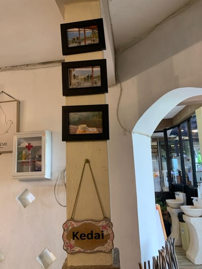 The image shows the interior of a building. There are three framed photos of outdoor scenes mounted on a vertical pillar. There is a sign that reads "Kedai" hanging on the pillar. There is a first aid kit hanging on the wall behind the pillar. An arched doorway on the right side of the image leads into an exterior space with tables and chairs. The location is in Kecamatan Borobudur, Indonesia, at latitude -7.606 and longitude 110.18. The scene appears to be in a restaurant or café.