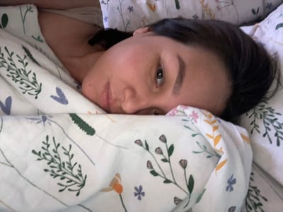 A woman lies in bed under a floral-patterned duvet. Her face is partially visible beneath the covers. The floral pattern includes green and purple motifs. The woman has dark hair and appears to be resting or sleeping. There is no information to place the location as Verkh-Katunskoye, Russia.