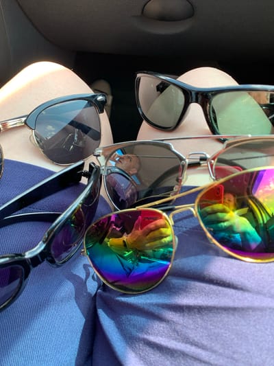 A close-up, overhead shot shows several pairs of sunglasses on a person's lap, likely within a vehicle. The sunglasses are in various styles, with some featuring mirrored, rainbow-colored lenses that reflect a person taking the photo holding a red smartphone. The background is dark and out of focus, possibly the interior of a car. The person's legs and a portion of a blue garment are visible. There are no recognizable landmarks or indications of the location being in Азерскі сельскі Савет, Belarus.