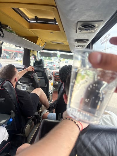 The image depicts the interior of a passenger bus or shuttle, viewed from a first-person perspective looking forward and slightly to the right. In the immediate foreground, a hand, adorned with black and red string bracelets, holds a clear plastic cup containing a transparent, effervescent liquid.
In the midground, a male passenger with short dark hair, wearing a black t-shirt and black shorts, occupies a black leatherette seat. His right arm, bearing a red string bracelet and a silver chain bracelet, rests on the seatback. A clear plastic cup with liquid sits in a holder on his seat. His left leg, clad in black athletic shoes, is extended. A blue plastic water bottle is visible in a mesh pocket on the back of the seat ahead of him. Further forward, the back of another person's head is visible in the vehicle's cockpit area.
The bus interior features a yellow fabric ceiling, overhead luggage compartments, and integrated air vents and lights. Through the front and side windows, an urban street scene is visible. On the left, a building displays a partial sign reading "FABRICA DE C...". A street with multiple vehicles, including cars parked and in motion, is visible, bordered by trees and other buildings. On the upper right window of the bus, the text "SALIDA DE EMERGENCIA" (Spanish for Emergency Exit) is visible, which contradicts the provided location of Jilava, Romania, suggesting a Spanish-speaking region.