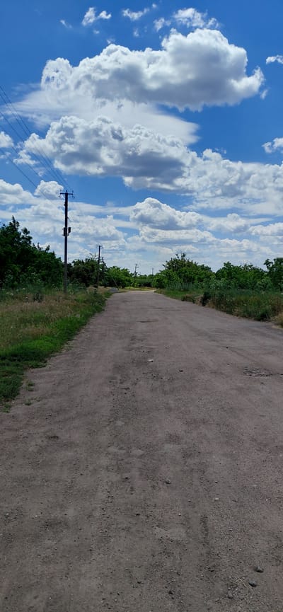 The image depicts a straight, unpaved road with a rough, dirt surface, stretching into the distance. The road is bordered on both sides by lush green vegetation, including trees and grass. Several utility poles with wires are visible along the left side of the road, indicating the presence of infrastructure. In the background, the road appears to curve slightly to the right, and more trees and vegetation can be seen. The sky above is a vibrant blue, populated with large, fluffy white clouds. The overall impression is a rural or semi-rural scene, likely in a countryside setting. The presence of utility poles and the road suggest it is likely a pathway within or leading to a populated area, possibly the outskirts of Ketrysanivka, Ukraine, given the prompt's context. No people or vehicles are present in the image.