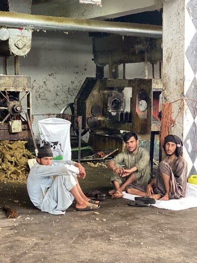 Three men sit on a white cloth in what appears to be an industrial setting in Shurandam, Afghanistan. One man is seated with his arms wrapped around his legs, and he wears a black beanie and a light-colored outfit. The second man sits cross-legged, with his hands clasped together, wearing a greenish-brown shirt. The third man, wearing a headscarf and dark clothes, also sits cross-legged. Scattered on the floor are sandals and small pieces of debris. Behind the men, industrial equipment is visible, including a large metal machine, ductwork running along the ceiling, and a white bag with a cow illustration. The walls are stained and appear to be in disrepair. A checkered pattern is also seen on a pillar.