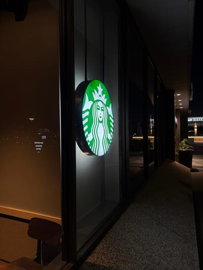 The image shows a Starbucks store in Maryland Heights, United States of America, at night. A large, illuminated Starbucks logo is visible on the exterior of the building, featuring the iconic siren. The logo is a circular sign with a green background and a white siren. The store has large glass windows, through which some interior lighting can be seen. Reflections of the exterior environment, including streetlights and some text, are visible on the glass. To the right of the entrance, there is a paved walkway and some planters. The overall scene suggests a well-lit commercial area at nighttime.