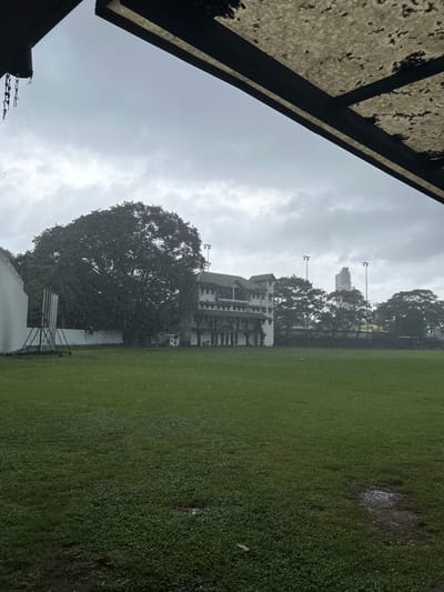 An outdoor scene captures a cloudy, rainy day at a cricket ground in Colombo, Sri Lanka. The foreground is dominated by a lush green field, partially wet from the rain, with visible puddles on the surface. In the midground, a large, mature tree stands prominently to the left, its dark foliage a stark contrast to the lighter sky. To the right of the tree, a multi-story building with balconies and a white facade is visible. Beyond the building and the tree, several other trees are scattered across the landscape. Two tall light poles with lamps stand in the distance, illuminated against the overcast sky. A modern, tall building rises in the far background. Overhead, the edge of a roof structure with chains hanging from it is visible in the upper portion of the frame. The rain is evident as streaks of white against the sky and scattered droplets on the ground and in the air. No people are visible in the frame.