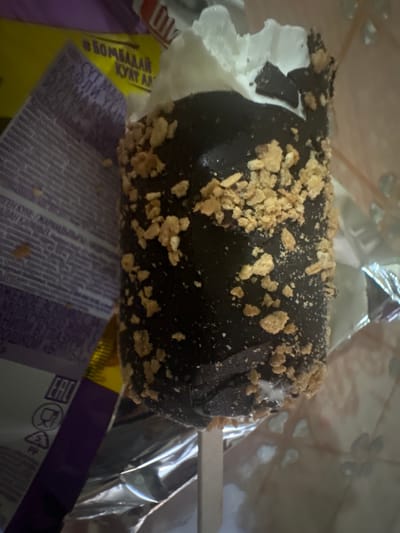 The image shows a close-up of a chocolate-covered ice cream bar on a stick, partially unwrapped. The ice cream is coated in dark chocolate and sprinkled with small, crumbly pieces, possibly nuts or cookie bits. A portion of the white ice cream is visible on the left side where the chocolate coating has broken away. The ice cream appears to be resting on its wrapper, which is made of shiny, crinkled material, likely foil. In the background and to the left of the ice cream, parts of a yellow and purple wrapper are visible with Cyrillic text printed on them. One section of the yellow wrapper has the text "• БОМБАДАЙ КУAT AA". Another section of purple wrapper has text that is partially legible, including what appears to be "СҮТ МАЙ" and "ВАФЛИ ҮСІН". The setting seems to be indoors, likely a kitchen or a place where food is prepared or consumed, given the presence of food wrappers and the focus on the ice cream itself. There are no people visible in the image. The lighting is somewhat dim and uneven, suggesting it might be taken in natural light or with indoor lighting. The overall impression is a casual, close-up shot of a sweet treat.