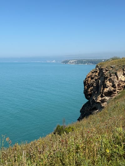 The image depicts a coastal landscape in Balgarevo, Bulgaria. A rocky cliff covered with greenery overlooks the blue sea. The sky is clear and bright. In the distance, a stretch of land is visible along the horizon, appearing hazy. There are no people or visible interactions in the frame. The focus is on the natural scenery.