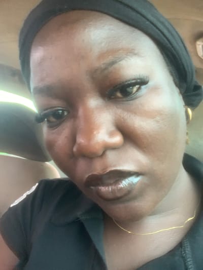 This is a close-up, low-angle selfie of a person's face, taken outdoors during the daytime. The person has dark skin, is wearing a black head covering, and has makeup on, including mascara and lip gloss. A thin gold chain is visible around their neck. The lighting suggests it is a sunny day, with bright light illuminating the scene from the side and below. The background is blurred, but appears to be outdoors, possibly in a vehicle given the presence of what looks like a car seat. The overall mood is casual and personal. There is no visible text in the image.