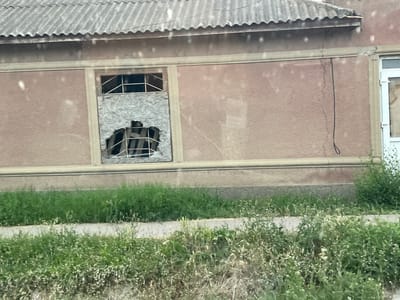 This media file shows an exterior view of a dilapidated building in Congaz, Moldova. The building has a corrugated metal roof and a textured, light brown stucco exterior. A prominent feature is a broken window, which has been boarded up with plywood. The plywood is damaged, revealing a metal grate behind it. The window frame is made of metal. A dark electrical wire hangs down the side of the building. A crack is visible in the stucco near the top of the building. To the right of the broken window, another window is partially visible, with a white frame and what appears to be a door or window covering inside. The building is bordered by a strip of overgrown green grass and weeds, suggesting neglect. A lighter-colored concrete pathway or curb runs in front of the overgrown vegetation. The scene suggests a rural or semi-abandoned location. The lighting is soft, indicating it might be daytime, but the overcast sky or time of day is not definitively clear. There are no people or animals visible in the frame. The overall mood is one of decay and abandonment. No visible text is present.