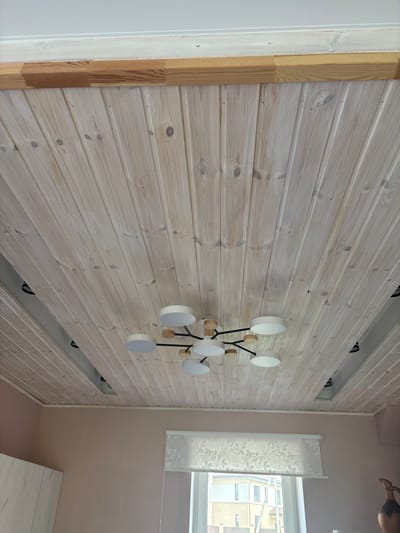 The image shows a wooden-paneled ceiling in a room. The wood planks are oriented lengthwise and have a whitewashed or lightly stained finish, revealing the grain and knots. A modern chandelier with multiple white circular lamps and wooden accents hangs from the center of the ceiling. In the background, the top of a window with a sheer patterned curtain is visible, along with a portion of a light pink wall and a part of a light-colored cabinet or wardrobe. The ceiling appears to be part of an interior space, likely a residential setting, possibly in the city of Dolce Vita KP, Russia, given the context.