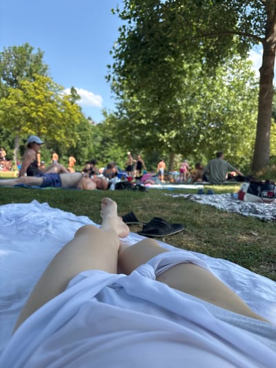 The image depicts a sunny day at a public park or recreational area, likely near Bürgstadt, Germany, given the user's prompt. In the foreground, a person's legs are visible, lying on a white blanket on a grassy area. They are wearing a white garment that drapes over their legs. A pair of black sandals are positioned to the left of the blanket.

In the background, several people are scattered across the grass, some lying down and others sitting. They appear to be enjoying the outdoors, possibly sunbathing or picnicking. Trees provide shade in various parts of the scene, and the sky is clear blue with a few wispy clouds. The overall atmosphere suggests a relaxed, casual outdoor gathering. The presence of trees and the general ambiance are consistent with parks found in or around Bürgstadt, which is known for its natural beauty and outdoor activities.