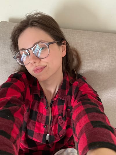 A young adult female is depicted in a close-up, high-angle selfie, identified as being in Scafati, Italy. She has fair skin, brown hair, and wears round, clear-framed eyeglasses with dark temples, which show reflections of what appears to be a window. Her lips are slightly pursed, forming a subtle smile. She is wearing a red and black buffalo plaid shirt or pajama top; the top two buttons are unfastened, revealing a small amount of white fabric underneath. The individual is positioned against a light beige or off-white fabric-covered headboard or upholstered backrest, with a plain light-colored wall visible in the upper background.