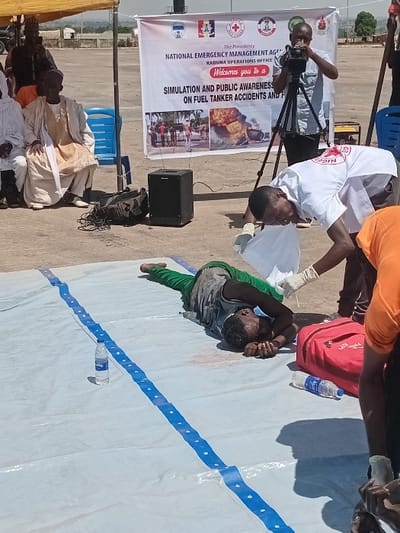 A man lies on a white tarp in a training exercise for a simulated fuel tanker accident in Katabu, Nigeria, at the coordinates 10.71 latitude and 7.514 longitude. A man in white, likely a first responder, attends to the man lying down, holding a white cloth near his head and wearing white gloves. Two bottles of water, a red medical bag and a blue stripe tarp are visible on the ground. In the background a cameraman films the event. A banner featuring the logo of the "National Emergency Management Agency", Kaduna Operations Office is in the background, along with several people watching the exercise. The exercise seems to be part of a public awareness campaign. The environment is sunny with a large canopy tent and chairs in the background. The cameraman is wearing a blue shirt and black pants. The cameraman is holding a video camera on a tripod. The man lying on the tarp is wearing a green shirt and a brown pants. The first responder is wearing a white shirt and black pants. The people in the background are wearing a variety of clothing.