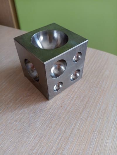 The image shows a metal cube with hemispherical indentations on its faces, placed on a light-colored wooden surface. The cube is positioned at an angle, with one face showing a large concave depression and two adjacent faces each displaying multiple smaller concave depressions of varying sizes. The surface it rests on has a wood grain texture. In the background, a green wall is visible. The scene appears to be indoors, likely on a table. No people or actions are depicted. The object is a forming or dapping block, a tool used in jewelry making and metalworking. The image does not contain information about the city of Vilyuysk, Russia.