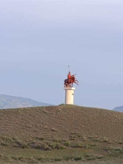 The image depicts a statue of two horsemen atop a cylindrical pedestal on a grassy hill. The statue appears to be made of bronze or a similar material with a reddish-brown patina. The horsemen are clad in what appear to be historical or traditional attire, one holding a spear. The pedestal is light-colored and shows some texture, possibly concrete. The hill is covered in dry grass and sparse vegetation, with undulating terrain. In the background, rolling hills and mountains are visible under a clear, pale blue sky. The overall scene suggests an outdoor monument in a natural landscape, likely in a rural or semi-rural setting within the city of Kyzyl-Jyldyz, Kyrgyzstan.