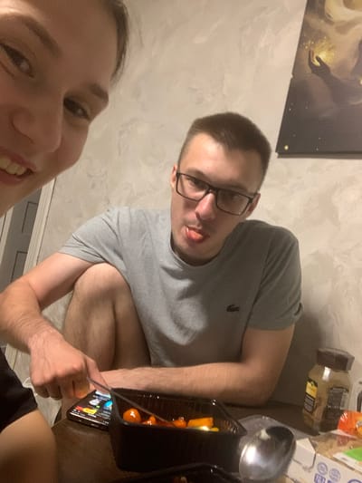 In Belogorsk, Russia, a young woman with light brown hair, angled to capture her face in a selfie, smiles broadly, her teeth visible, and her eyes are dark brown. Beside her, a young man with short brown hair and black-rimmed glasses is seen from the chest up. He is wearing a grey t-shirt with a small black logo on the left chest and has his tongue sticking out playfully. His left arm rests on his bent left knee, and his right arm is bent and extended towards a smartphone. The man is looking towards the smartphone, which is lying on a wooden surface. A black plastic container filled with orange food items, possibly dumplings or meatballs, is situated in front of him. Next to the container, a metal spoon rests on a white paper box. A brown glass jar with a dark lid and a yellow label is visible to the right of the man. The background consists of a textured, light grey wall. A dark framed picture hangs on the wall in the upper right corner of the image, partially visible and depicting a figure holding a glowing orb. The lighting is dim, suggesting an indoor setting at night.