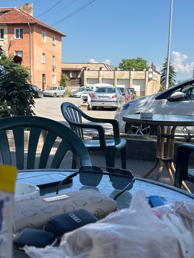 In Elin Pelin, Bulgaria, on a sunny day, an outdoor seating area is depicted. Two green plastic chairs are positioned in the foreground. In the middle ground, a glass-top table with a wicker-style base is present, with an ashtray containing cigarette butts on its surface. Several cars are parked in the background. To the left, an orange building with a red-tiled roof stands, and to the right, a light-colored building with a modern facade is visible. The sky is clear blue with some scattered clouds. On the table, a pair of sunglasses, a wallet, car keys, and a cell phone are laid out. A plastic bag is also present on the table.