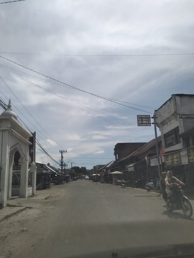 The image depicts a street scene in the city of Pulorayeuk, Indonesia, under a cloudy sky. The road, made of asphalt and dirt, runs through a built-up area with buildings lining both sides. On the left, a white structure with arched entrances and a domed roof, possibly part of a mosque or religious building, stands prominently. Adjacent to it, a dark billboard is visible.
The street is flanked by various establishments, including shops with awnings and tiled roofs. Several motorcycles are parked or in motion. In the foreground on the right, two individuals on a motorcycle are captured in motion, with the rider wearing a light-colored headscarf and the passenger behind them also wearing a headscarf. Other motorcycles, including a light blue scooter, are visible in the mid-ground.
Utility poles with a tangle of electrical wires crisscross the sky above the street. A traffic signpost with a square sign featuring a grid pattern is mounted on a pole on the right side of the road. The overall environment suggests a moderately populated urban or semi-urban area with ongoing daily activities. The buildings appear to be a mix of older and newer structures, some showing signs of wear.