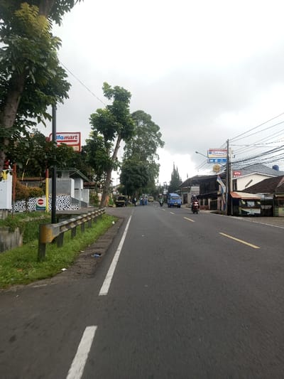 The scene depicts a street view on an overcast day in Kandapola, Sri Lanka, looking down a paved two-lane road. The sky is uniformly grey with heavy cloud cover.
The asphalt road, dark in color, extends into the distance, with a solid white line marking its left edge and a dashed yellow line indicating the center. On the immediate left, a grassy shoulder runs alongside the road, bordered by a yellow-topped, black metal guardrail. Below the guardrail, a concrete drainage channel is visible, with some litter present.
Beyond the guardrail on the left, an area features several large green trees. A white building with a patterned black and white section is visible, adorned with a red circular sign featuring white text that reads "KELUAR." Further down, a prominent red and white "Alfamart" sign is affixed to a building, partially obscured by foliage.
On the right side of the road, a dense network of utility poles and overhead power lines runs parallel to the street. A multi-layered blue and white sign for "Indomaret" is clearly visible, with additional smaller signs below it. Adjacent to this, a building displays an "ATM" sign. Various single and multi-story buildings, some with shaded awnings or stalls, line this side of the street, suggesting commercial activity.
Traffic includes a red motorcycle with a rider wearing a helmet, moving away from the viewer on the right side of the road. Further in the distance, a blue van or minibus also travels away. Several other vehicles, including a dark pickup truck and other motorcycles, are visible further down the road, either stationary or moving slowly. No pedestrians are distinctly visible. The environment combines natural elements like trees with urban infrastructure and commercial establishments.