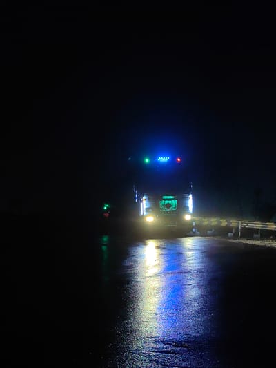 A truck is stopped on a wet road at night. The truck has blue and green lights on its roof and red and yellow lights on its front. The road reflects the lights. There are guardrails on the side of the road. The scene appears to be in Kulang, India.