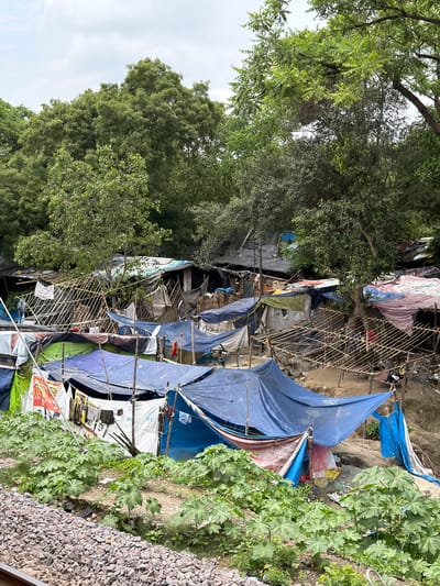 In Shuklaganj, India, a dense cluster of makeshift dwellings, constructed primarily from tarps and bamboo, occupies a sloped area bordered by a railway track with gravel ballast. The shelters are of varying sizes and colors, predominantly blue, green, white, and red, with some featuring additional materials like corrugated metal sheets and fabric. They are supported by bamboo poles and ropes, creating a temporary and precarious settlement. Vegetation, including large-leafed plants, grows in front of and around the dwellings, indicating a possible undeveloped or informal area. The sky above is overcast with clouds. There is no discernible human activity or interaction in the image. The environment appears to be a mix of natural growth and informal human settlement.