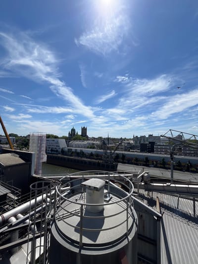 The image captures an urban waterfront scene under a bright blue sky with wispy clouds. Several birds are in flight against the expansive sky, some appearing as small silhouettes. In the foreground, a large industrial reel or spool made of metal stands prominently, casting a distinct shadow. Surrounding it are various metal pipes and structural elements, suggesting an industrial or port area.
Beyond the immediate industrial foreground, a city skyline unfolds along the waterfront. Buildings of varying heights and architectural styles are visible, with some featuring green roofs or lush trees interspersed. A church with a prominent spire and another building with a dome are notable landmarks in the cityscape. The water of a canal or river runs alongside the buildings. Construction scaffolding is visible on one of the buildings, indicating ongoing development or renovation.
The sunlight suggests it is daytime, likely late morning or early afternoon given the brightness and angle of shadows. The weather appears clear and sunny. The presence of industrial structures, the waterfront, and the specific architectural style of the buildings in the background are clues to the location. The overall mood is one of a bustling, active urban environment bathed in sunlight. No visible text is present in the image.