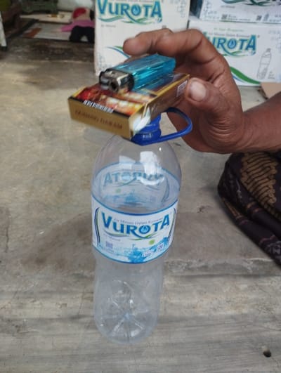 A person's right hand holds a blue disposable lighter and a brown and gold cigarette pack, which are resting on the mouth of a clear plastic water bottle. The scene is set on a light gray concrete floor, indicating an indoor or sheltered outdoor location in Kecamatan Panji, Indonesia.
The primary subjects are the hand, the lighter, the cigarette pack, and the water bottle. The water bottle is mostly empty, containing only a small amount of liquid at the bottom. It has a blue and white label. The cigarette pack is predominantly brown and gold with some red detailing. The lighter is transparent blue. The hand appears to be that of an adult.
In the blurred background, several white cardboard boxes are stacked, and a piece of dark, intricately patterned fabric is visible on the right side of the frame, possibly a local textile like a sarong.
The visible text includes "VUROTA" prominently displayed in blue on the water bottle's label, with "Air Minum Dalam Kemasan" (Packaged Drinking Water) also visible below it. The "VUROTA" brand is also discernible on the cardboard boxes in the background. Text on the cigarette pack is partially visible, appearing to be Indonesian, but not fully legible. The activity suggests the individual is either preparing to smoke or has momentarily placed their smoking items on the bottle. No specific time of day or weather conditions are discernible, though the lighting suggests daytime. No particular emotions are evident.