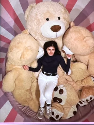 A woman is lying on a large pile of stuffed teddy bears. She is wearing a dark long-sleeved shirt, white pants, and sneakers. The teddy bears are of various sizes and are mostly beige in color. Some of the teddy bears have brown patches on their ears and paws. The scene is set on a floor with a striped pattern of pink and purple. The lighting suggests it is daytime. The woman appears relaxed and comfortable as she lies amongst the toys. There are no visible text elements in the image. The overall impression is one of playfulness and comfort, possibly in a home setting in Nijmegen, Netherlands.
