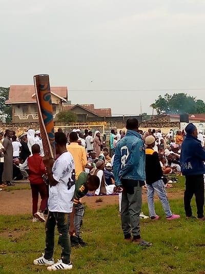 The image shows a gathering of people in an open field in Beni, Democratic Republic of Congo, located at coordinates 0.489 latitude and 29.464 longitude.  The ground is mostly covered with grass, and there are a few buildings in the background. There are approximately 50 people in the scene, most of them are men and boys. They are all dressed in casual clothing. Many are sitting on the ground, while others are standing.  A young man is standing in the foreground, holding a rolled-up rug or carpet.  There are several people standing behind him, looking at the rug or the camera. The people in the background are blurred, and it is difficult to tell what they are doing. There appears to be a small amount of smoke in the background. The scene is peaceful and there is no evidence of any activity that could be considered dangerous or harmful.