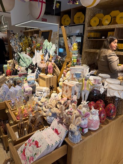 This image depicts the interior of a confectionery or gourmet food shop located in Felgueiras, Portugal, densely arranged with Easter-themed merchandise.
**People:** One female individual is visible on the right side of the frame, standing behind a display counter. She has dark hair and wears a light brown, long-sleeved top. Her head is turned to her right, and her hands are partially visible, suggesting she is attending to items on the counter. A second person, largely obscured, is partially visible on the far left, facing away from the camera.
**Subjects/Objects:** The central focus is a large, multi-level display counter overflowing with Easter products. This includes numerous plush rabbit toys, many dressed in checkered blue and white fabric, and various ceramic rabbit figurines, some holding miniature teacups. Large, foil-wrapped chocolate eggs in gold, silver, and green are interspersed throughout the display, alongside smaller gift-wrapped items. Several decorative red and black patterned Easter eggs are visible on the right side of the display, adjacent to two large glass jars containing chocolate-covered nuts or similar confectionery. In the foreground on the left, wooden display drawers hold packaged sweets, including white boxes with pink floral designs labeled "Delfin Chocolate." A tall, stacked display of yellow ceramic teacups and a teapot, topped with a rabbit figurine, stands on a wooden easel-like structure. In the background on the right, wooden shelves display octagonal yellow boxes, some branded "BOCCA." A large, white egg-shaped decoration with red stripes hangs from the ceiling.
**Actions/Interactions:** The female individual appears to be working, possibly organizing or serving from the counter. No direct interaction between individuals is observed.
**Environment:** The interior space is brightly lit by ceiling fixtures, including a long rectangular LED light. The decor incorporates extensive use of wood for shelving and display units. The abundance of rabbit motifs, decorative eggs, and themed packaging indicates a festive, seasonal retail environment focused on Easter.