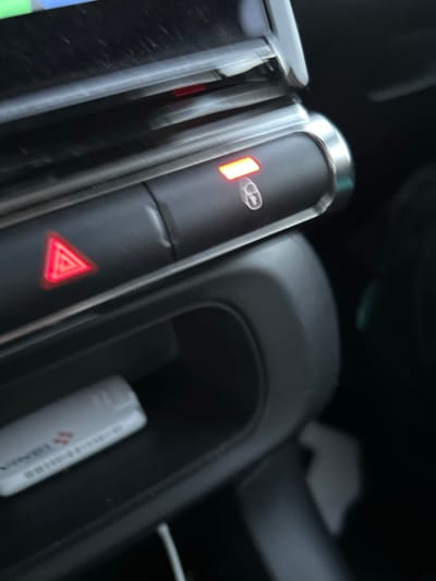 A blurry, close-up shot inside a car focuses on the central console. The center console's buttons are visible; the top button has a person icon and a horizontal bar, lit in orange. Below is a hazard light button, lit red with a triangle icon. A compartment to the right holds a small, white tube, potentially lip balm or similar. The surrounding interior is dark and out of focus.
