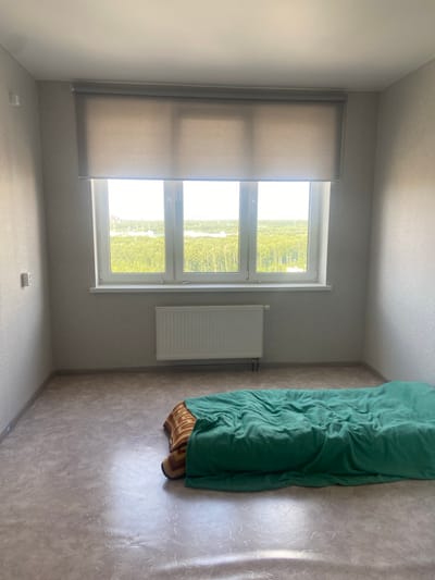 The image shows a sparsely furnished room with a large window. The room has light grey textured wallpaper and a greyish-beige floor with a swirling pattern. A horizontal white radiator is mounted below the window. A light-colored roller blind partially covers the window, which overlooks a green landscape with trees and some buildings in the distance. In the foreground, a mattress covered with a crumpled green blanket and a striped blanket is placed directly on the floor. There is a white electrical outlet on the left wall. The scene appears to be an interior of a residential building in Kazan, Russia.