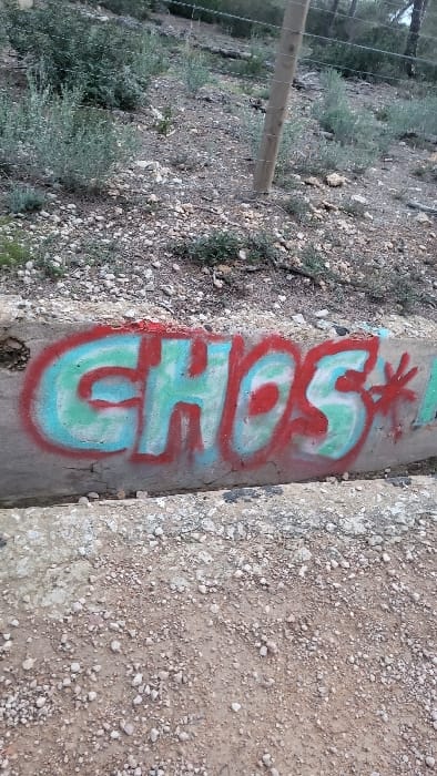 This image depicts a segment of a natural, undeveloped outdoor area in Santa Ponça, Spain. The foreground consists of a dirt and gravel ground surface. Immediately above this, a weathered concrete retaining wall or curb runs horizontally, displaying graffiti. The graffiti consists of the word "GHOS" rendered in light blue spray paint with a distinct red outline, followed by a red asterisk symbol. The upper midground reveals a gently sloping, unpaved terrain composed of reddish-brown soil and small stones, interspersed with sparse, light-colored scrub brush and low vegetation. A wooden fence post is visible in the upper right, secured into the slope, with several strands of wire (appearing to be fencing wire) attached horizontally. The background shows a more densely vegetated hillside with trees and shrubs. No individuals, animals, or vehicles are present.