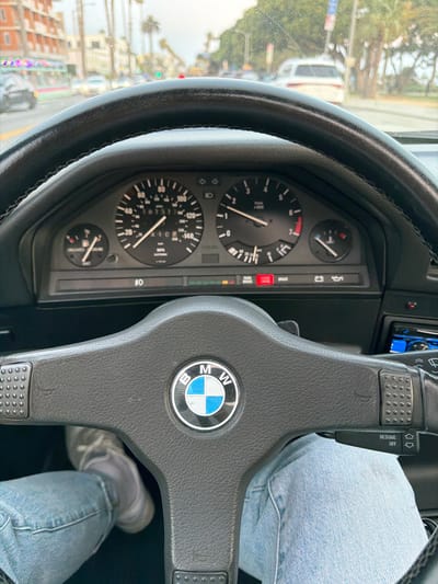 The image is a first-person perspective from inside a BMW, looking at the dashboard and the road ahead through the windshield. The driver's legs, clad in light blue jeans and grey athletic shoes, are visible in the lower part of the frame. The steering wheel, featuring the BMW logo, is prominent. The dashboard displays a speedometer, tachometer, fuel gauge, and temperature gauge. Several warning lights are illuminated, including "PARK BRAKE" and "BRAKE". The exterior view shows a street scene with buildings, palm trees, and other vehicles, suggesting an urban environment. The lighting and atmospheric conditions, with some moisture on the windshield, indicate it may be overcast or raining. The architecture of the buildings and the presence of palm trees are characteristic of a Southern California city like Santa Monica.