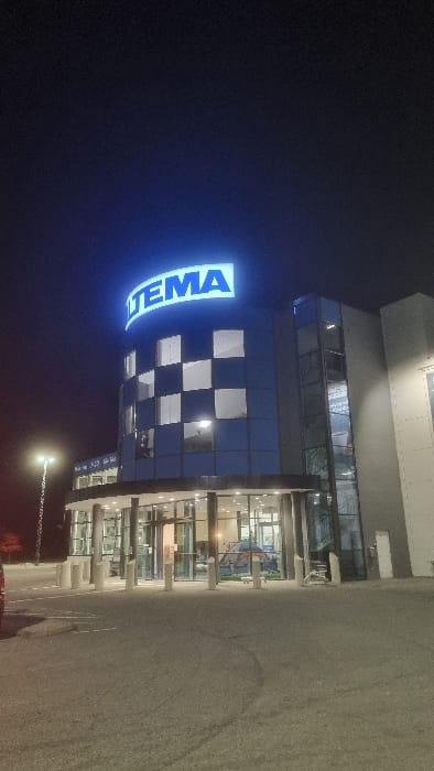 The image displays the exterior of a Jula retail store at night, specifically identified as the Jula Norrköping - Ingelsta location at Koppargatan 13, 602 23 Norrköping, Sweden. The multi-story commercial building features a prominent curved facade with an alternating pattern of dark blue panels and reflective windows. An illuminated blue sign bearing the name "JULA" is mounted prominently on the roofline. The ground level comprises a glass-enclosed entrance area supported by several grey cylindrical pillars. The interior is visibly lit, revealing merchandise such as a blue tent-like structure through the glass. Concrete bollards are positioned in front of the entrance. To the right of the curved section, the building extends with vertical glass panels and a lighter grey wall. The foreground consists of an asphalt parking lot, illuminated by the building's lights and a tall, multi-lamp streetlight positioned on the left side of the frame. The rear quarter panel of a red vehicle is partially visible in the bottom left corner. The sky is dark blue, indicating nighttime. No individuals are present in the visible scene.