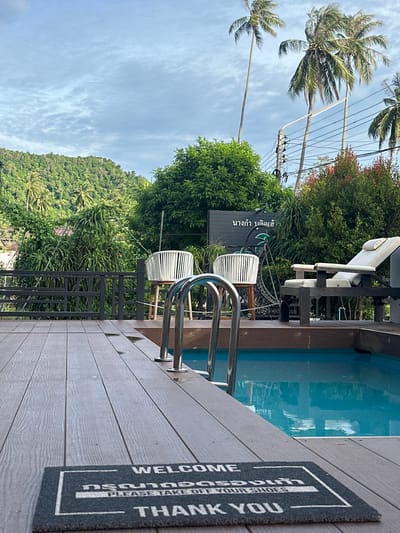 The image depicts a serene outdoor scene in Don Sak, Thailand, featuring a private swimming pool area. A wooden deck surrounds the pool, leading to a welcome mat that reads "WELCOME, PLEASE TAKE OFF YOUR SHOES, THANK YOU." Two white woven chairs and a lounge chair are visible on the deck, suggesting a place for relaxation. Lush green vegetation, including palm trees and dense foliage, frames the scene, with a tropical hillside in the background. The sky is overcast with scattered clouds. A metal ladder descends into the clear blue water of the pool. The overall impression is one of a tranquil resort or private villa.