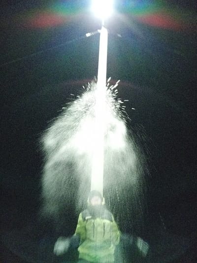 An outdoor night scene in Старый Шагирт, Russia, depicts a person standing beneath a brightly illuminated vertical pole. A significant volume of white liquid or snow particles is seen erupting and cascading outwards from the lower section of the pole, illuminated brightly by its light source. The person, positioned below the primary spray, wears a dark hooded head covering and a two-tone jacket, primarily light green/yellow with dark shoulders. Their arms are visible, slightly blurred due to motion, and they appear to be wearing dark gloves or mittens. The background is completely dark, devoid of discernible features.