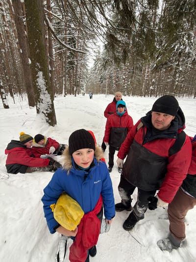 A winter scene in a snow-covered forest, identified as Люк, Russia. The image features a group of approximately eight individuals dressed in cold-weather gear. In the immediate foreground, a child, wearing a blue parka with a fur-trimmed hood and a black beanie, stands facing the camera, holding a yellow bag and a red garment. To their right, a man with a serious expression, wearing a red and black winter jacket, a black beanie, and a backpack, stands partially visible. Behind the child, on the left side of the frame, three individuals in red and black jackets are seated or partially reclined in the snow, one with a yellow and black beanie. Further down a cleared, snow-covered path that extends into the background, other individuals are visible walking or standing. All participants are clad in heavy winter clothing, primarily red and black jackets. The environment consists of dense evergreen trees heavily laden with snow, and the ground is entirely covered by a thick layer of fresh snow. The sky appears overcast.