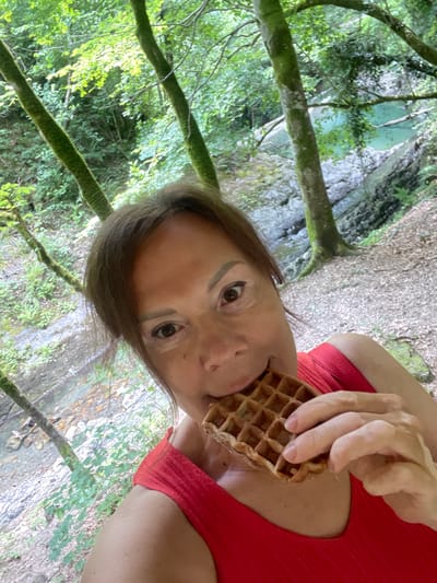 The image shows a woman in a red top holding a waffle to her mouth as she looks towards the camera. She is outdoors, with lush green foliage and trees framing the scene. A body of water, possibly a river or stream, is visible in the background, with rocky terrain and more trees. The lighting suggests it is daytime, and the weather appears to be pleasant, given the verdant surroundings. The woman's expression is neutral, and she seems to be enjoying her snack amidst nature. The location is identified as Khostinskiy, Russia, which is consistent with the natural, forested environment depicted. There is no visible text in the image.