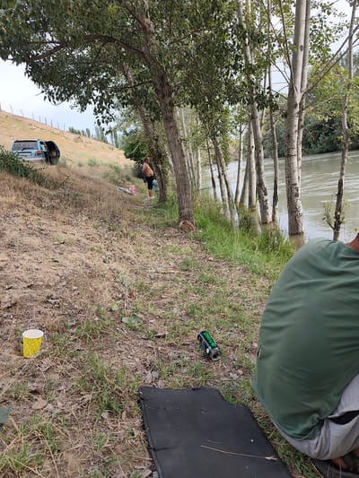 The image depicts a riverside scene in Ноошкен, Kyrgyzstan. A light blue minivan with its rear hatch open is parked on a dry, grassy slope. Near the vehicle, a man with his back to the camera is visible, wearing a green t-shirt and grey pants. In the foreground, a black mat is laid out on the ground, and a crushed can with "DRIVE" printed in green lettering lies beside it. To the left, a yellow paper cup with white polka dots rests on the dry grass.
The setting is a natural one, with a body of water on the right side of the frame and a line of trees, including tall, slender poplars, bordering the riverbank. The vegetation is dry and sparse in some areas, suggesting a warm season. The sky is overcast, indicating a cloudy or possibly hazy day. The overall atmosphere seems relaxed and casual, possibly indicating a picnic or a break by the river. The presence of the vehicle, mat, and cup suggests people are spending time outdoors, perhaps fishing or enjoying the scenery. The natural landscape with the river and trees is the dominant element, with the human presence being secondary and integrated into the environment.