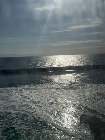 This image depicts a wide expanse of the ocean under a partly cloudy sky. The sun is visible and shining brightly, creating a strong reflection on the water's surface, which is choppy with visible waves and foam. In the distance, a faint silhouette of ships can be seen on the horizon. The sky is a mix of blue and grey clouds, with streaks of sunlight piercing through. The foreground shows the turbulent water closest to the viewer, with white foam and ripples. The scene evokes a sense of vastness and natural beauty. The location is identified as Unknown, Denmark.