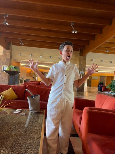 A young person with short, dark hair stands in a hotel lobby, making a face with their tongue sticking out and hands held up in a playful gesture. They are wearing a white short-sleeved button-up shirt and white pants with a textured pattern. To the left, a red sofa with a yellow pillow is visible, and in front of it, a wicker-topped coffee table holds a gray bag and some papers. To the right, another red sofa is partially visible. The background shows the hotel's interior architecture, including a wooden ceiling with exposed beams, stone pillars, and a staircase in the distance. The scene appears to be in a hotel lobby in Antigua, Spain, characterized by its warm lighting and comfortable seating areas.