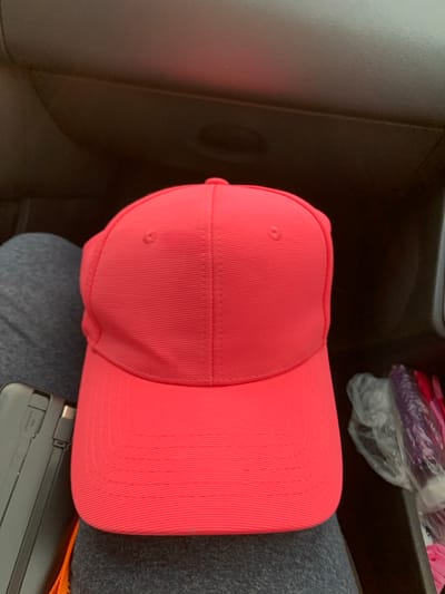 A close-up, overhead shot captures a bright coral pink baseball cap resting on a person's lap. The cap features subtle horizontal ribbing and a small button at the top. To the left of the cap, a black electronic device with a silver connector is visible. The background consists of dark, textured material, possibly the interior of a vehicle. To the right, a plastic bag contains purple and pink items, suggestive of toiletries or personal care products. The scene appears to be set in the rural area of Блеўчыцкі сельскі Савет, Belarus.