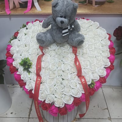 The image displays a large, heart-shaped arrangement of artificial roses, predominantly white with accents of red and pink along the outer edge. A grey teddy bear wearing a black and white striped scarf is seated on top of the roses. Red ribbons are wrapped around the arrangement, with the words "LOVE IS ETERNAL" printed in gold lettering.
The arrangement is placed on a surface that appears to be a shelf, with other decorative items and ribbons visible in the background. The overall presentation suggests a gift or a decorative piece for an event like Valentine's Day or an anniversary. The lighting is bright, indicating it was taken indoors during the daytime. The floor visible at the bottom of the image is tiled, suggesting an indoor commercial or domestic setting. No people are visible in the frame. The emotion conveyed is one of affection and romance, due to the heart shape and the inscription on the ribbons. The context of Urgench, Uzbekistan, places this scene in a specific geographical location, but this detail does not influence the visual description of the subjects or their arrangement.