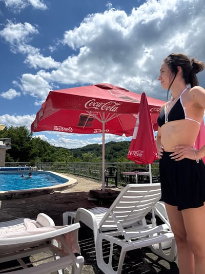 In Shipkovo, Bulgaria, on a sunny day with scattered clouds, a young woman stands by a swimming pool. She is wearing a black bikini top and black shorts, with her hands on her hips. A large red Coca-Cola umbrella is open behind her, and a smaller, closed red umbrella is next to it. A swimming pool with blue water is visible to the left, with two people swimming in it. White lounge chairs are scattered around the pool area. In the background, lush green trees cover rolling hills, and a building with a balcony is on the left.